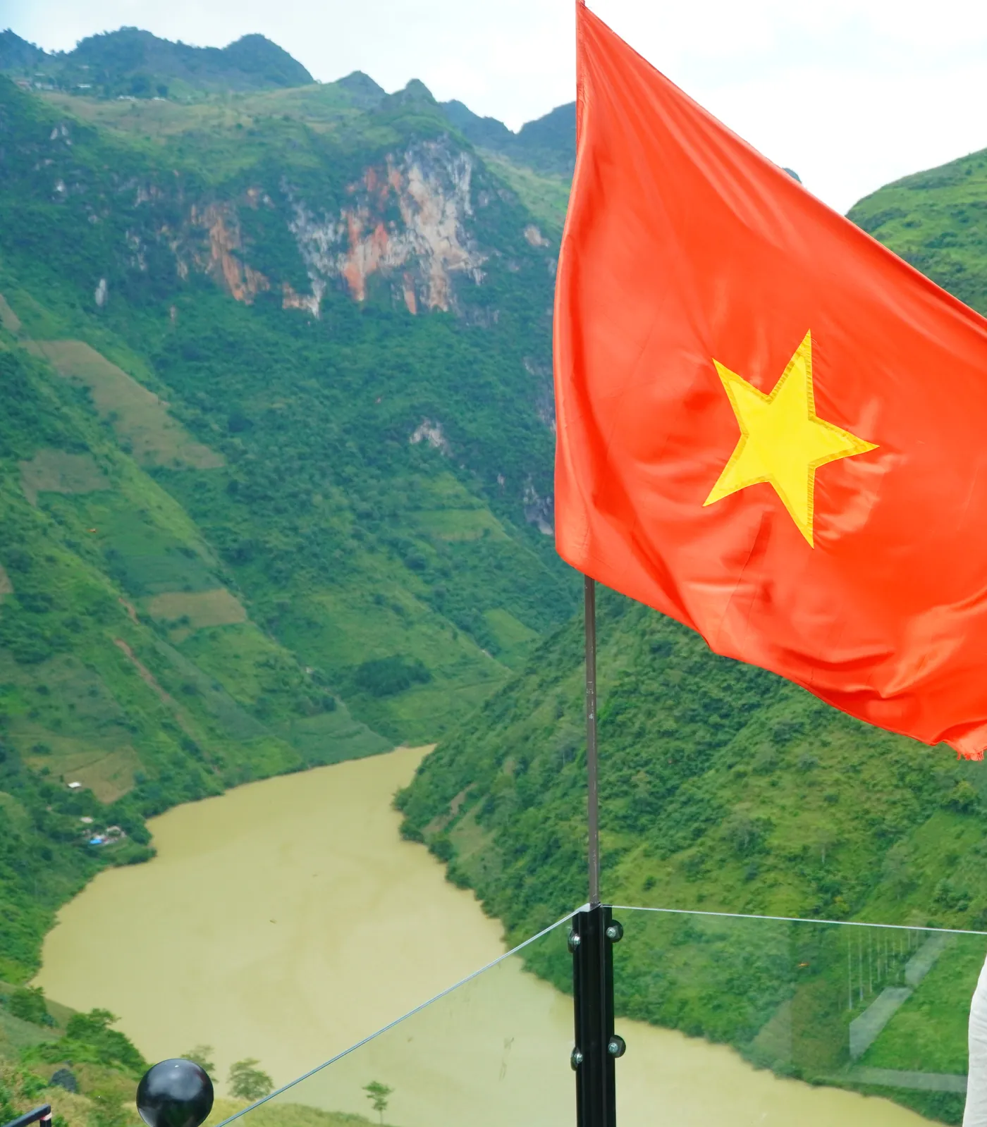 A Vietnamese flag on a pole at a viewpoint fitted with glass safety panels, with a wide bend of khaki-green river far below in a steep valley of green terraced slopes and grey limestone