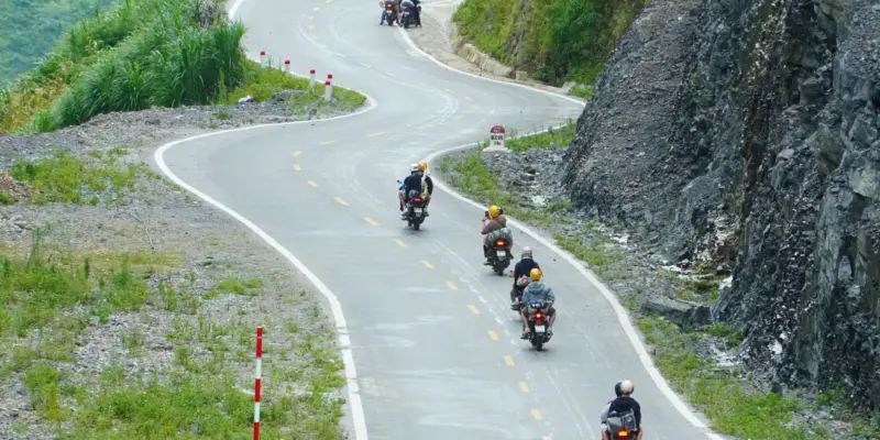 Motorbikes descending a newly surfaced two-lane mountain road that doubles back on itself, most strung out in a line and further machines riding abreast at the top of the frame where the road is cut off by the edge, every rider seen from behind and wearing a helmet, no faces in frame, with red and white marker posts along the left verge, a cut rock face on the right and a concrete drainage channel beside the carriageway