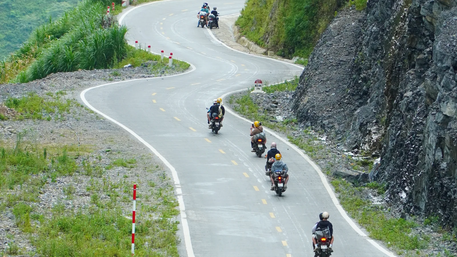 Motorbikes descending a newly surfaced two-lane mountain road that doubles back on itself, most strung out in a line and further machines riding abreast at the top of the frame where the road is cut off by the edge, every rider seen from behind and wearing a helmet, no faces in frame, with red and white marker posts along the left verge, a cut rock face on the right and a concrete drainage channel beside the carriageway