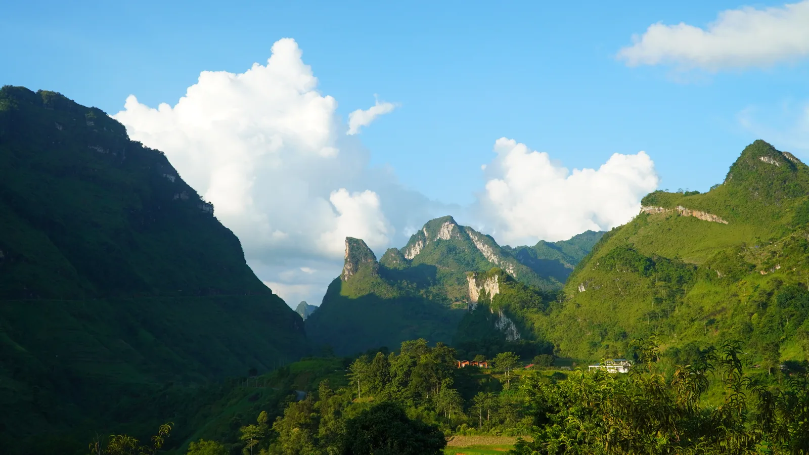 Limestone karst peaks under a blue sky with tall white cloud building behind them, a steep green ridge falling away on the left with a pale road line cut across its face, and a handful of red-roofed buildings among trees on the valley floor. No people and no vehicles are in the frame.