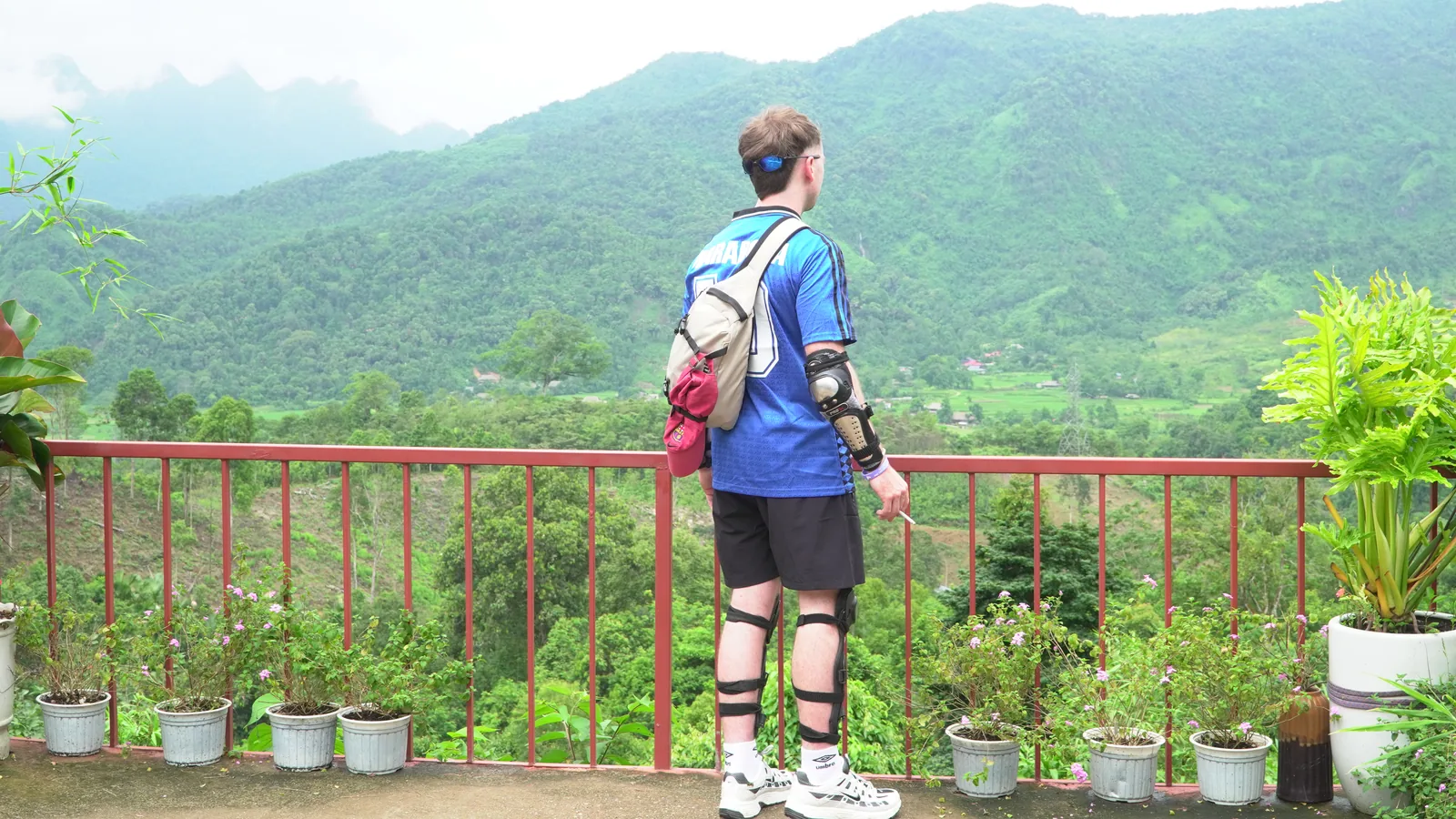 A rider photographed from behind at a viewpoint railing, wearing a blue football shirt and a sling bag, a hard elbow guard strapped to the right forearm and hinged knee armour strapped over both legs, looking out across a forested valley with cloud sitting on the far ridges and a scatter of buildings and green fields on the valley floor. The face is not in frame.