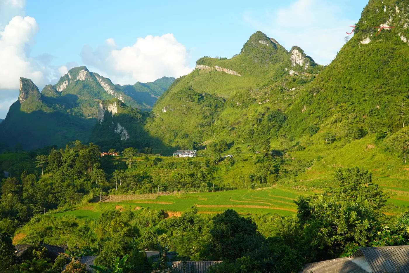 Grey limestone peaks rising in ranks under a bright overcast sky, their lower slopes cut into narrow cultivated terraces, and a thin pale track visible traversing one shoulder