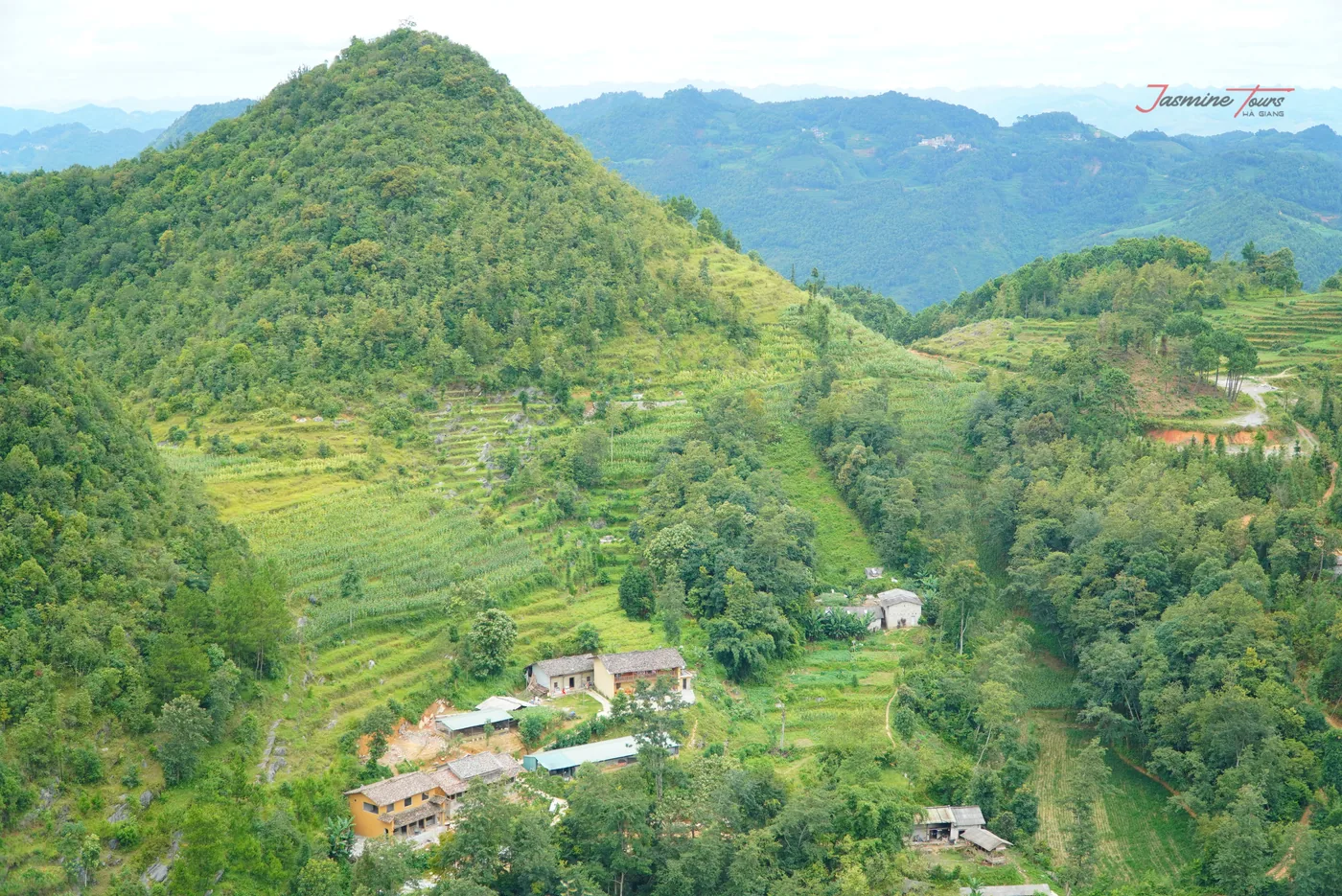 A steep green valley seen from a high road, cultivated terraces stepping up the near slope, a scatter of small buildings with pale roofs on the valley floor, and forested limestone ridges layered into haze behind