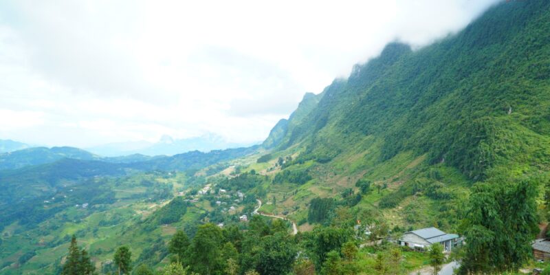 A wide green valley seen from high on the road, a settlement of small pale-roofed buildings strung along the valley floor and up the far slope, terraced fields cut into the hillsides, a single-storey roadside building at the right, and cloud sitting on the ridge above