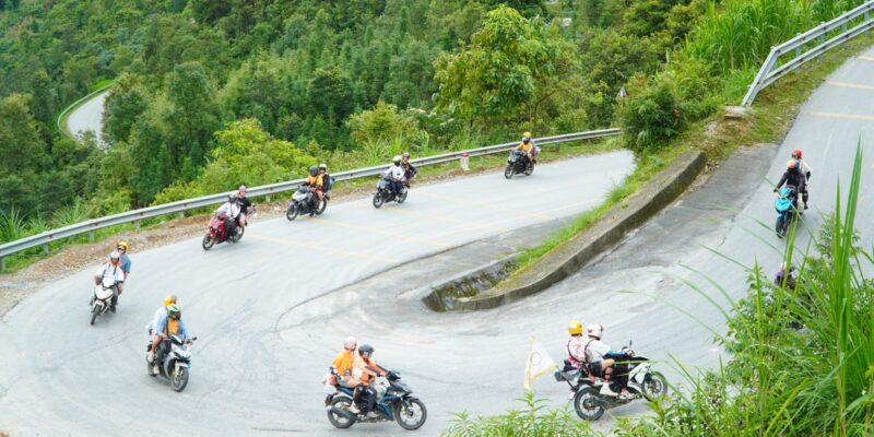 Looking down on a full hairpin bend where about nine small motorbikes are working through the turn at different points, several riders in orange jerseys, two carrying pillion passengers, one bike flying a small yellow pennant, a steel guardrail on the outside and dense green vegetation on the slope above