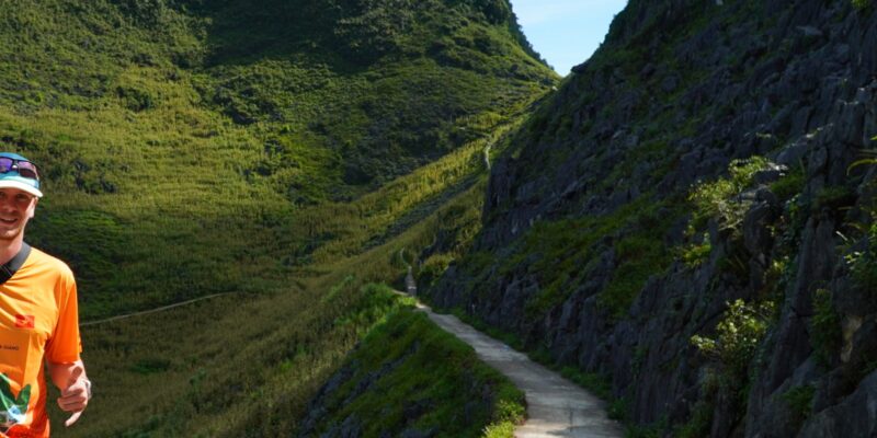 A narrow pale road running away between two steep green karst walls with a rocky verge on one side