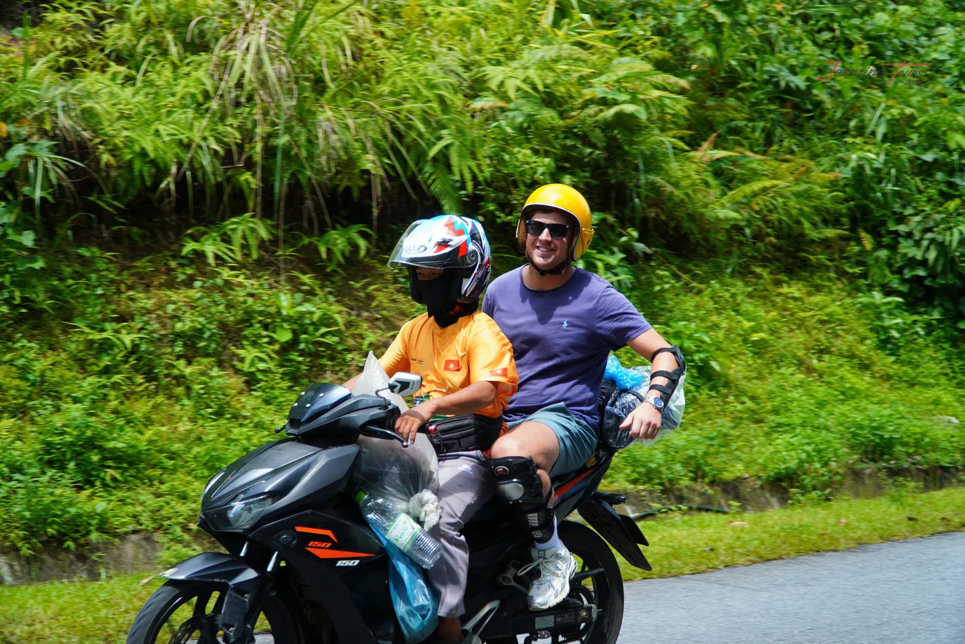 A guide and a passenger on a small motorbike on a two-lane road, the tarmac clean and dry with a concrete kerb along the uphill side, tall grass and dense green vegetation banked above them