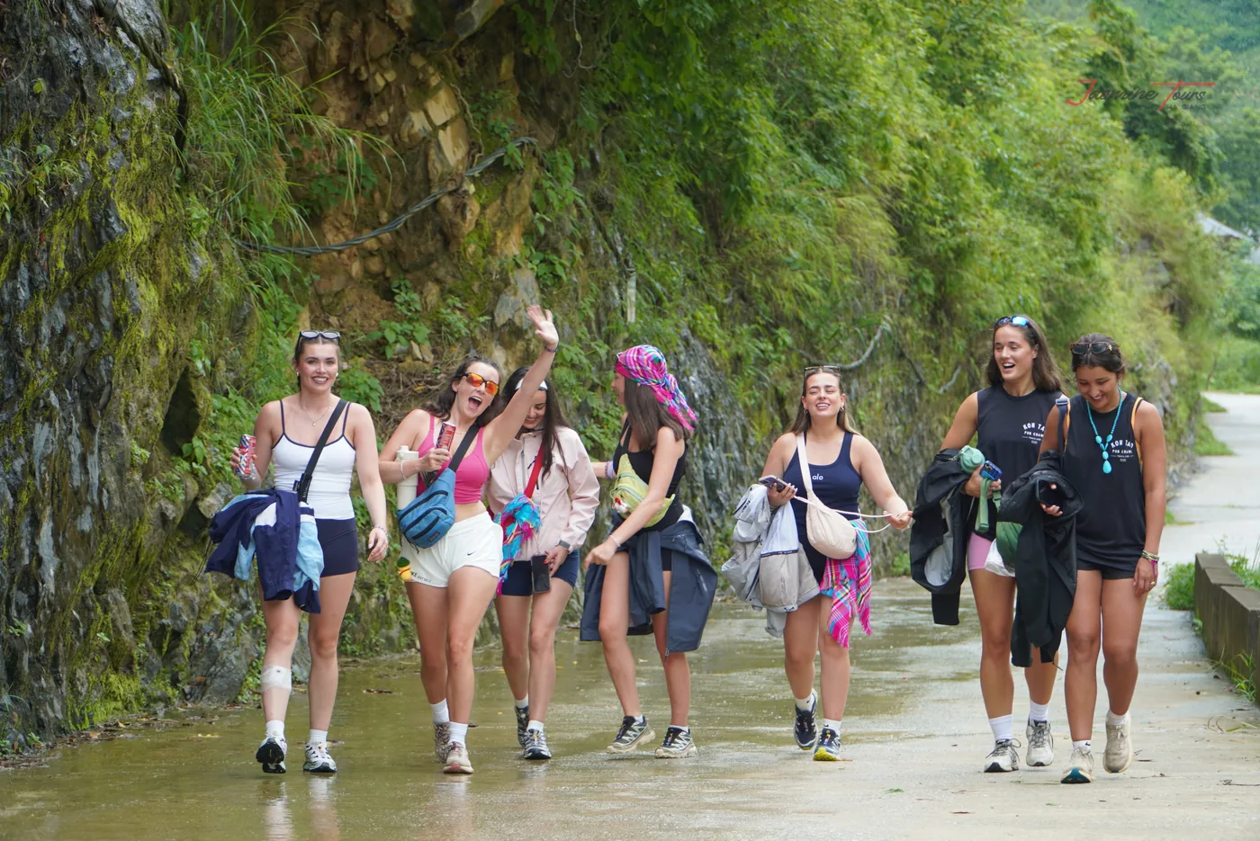 A group of travellers walking along a wet road below a rock cut, water lying across the carriageway and dark stains down the cut face, with green vegetation overhanging from above