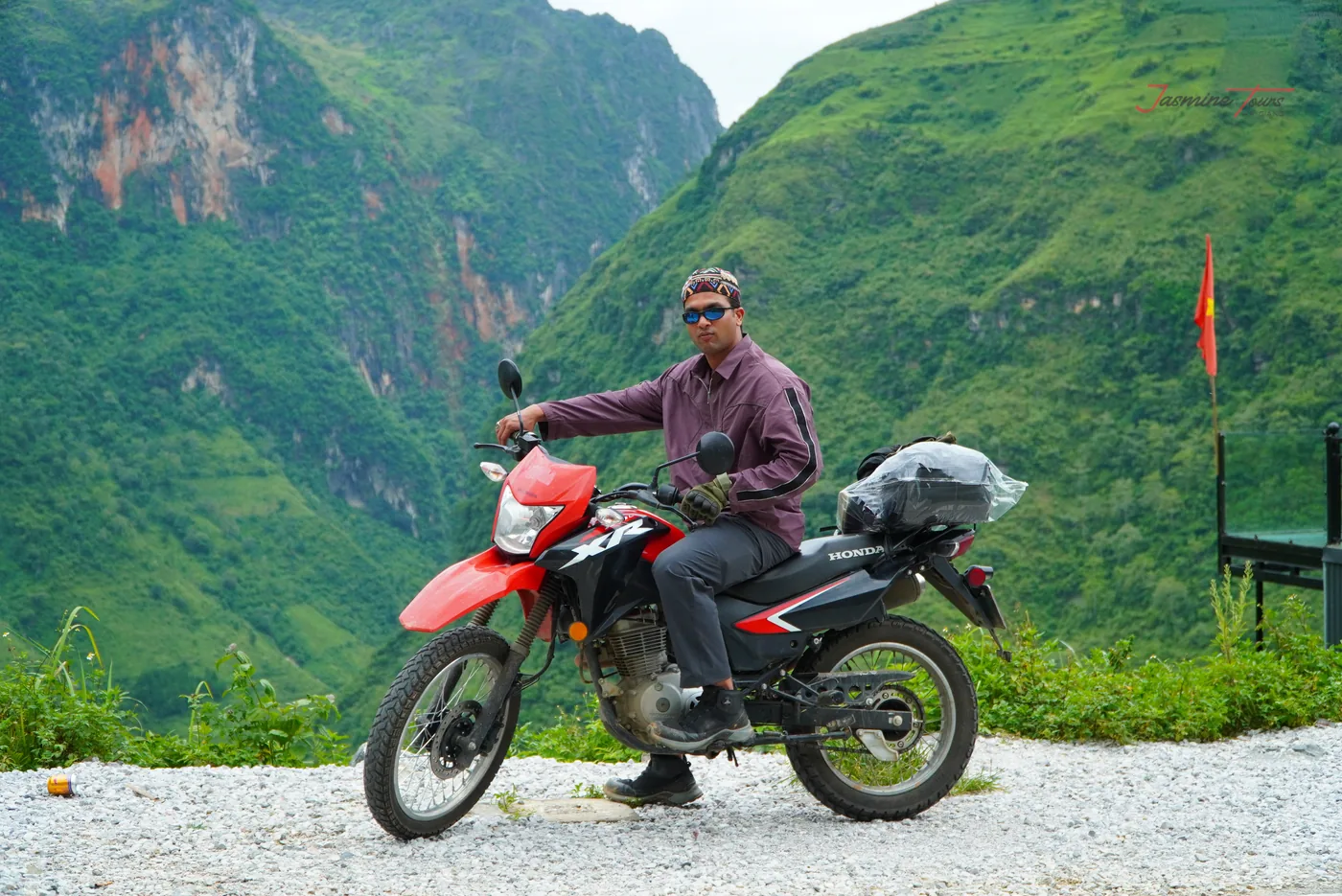 A man in a purple jacket sitting side-on on a red trail motorbike parked on a gravel shoulder, a rain-covered bag strapped to the rear rack, a red flag on a pole behind him and a deep green gorge falling away below