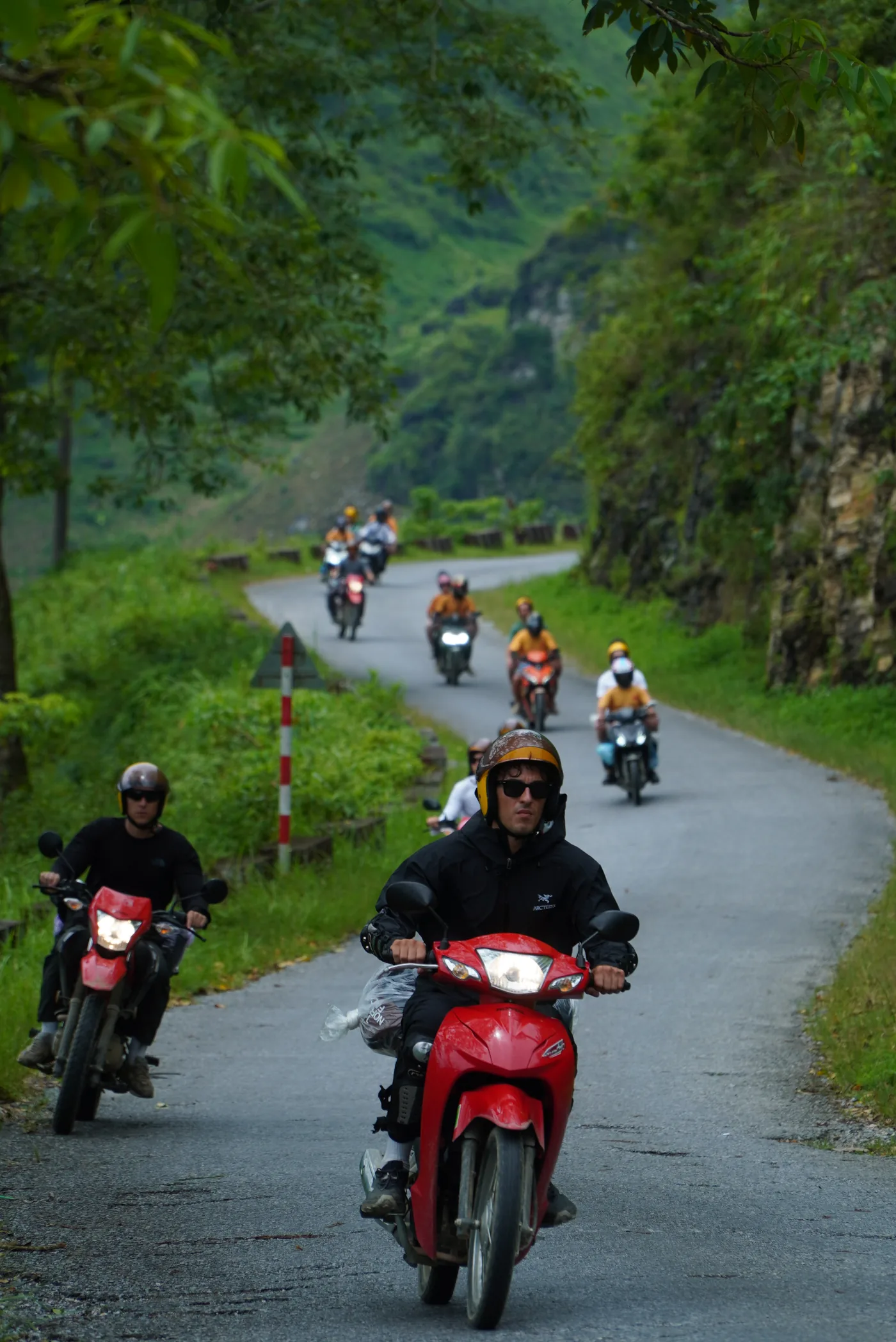 A column of riders coming down a narrow sealed road between a rock cut and a green verge, the nearest rider on a red semi-automatic in a dark jacket and sunglasses, red-and-white marker posts along the edge