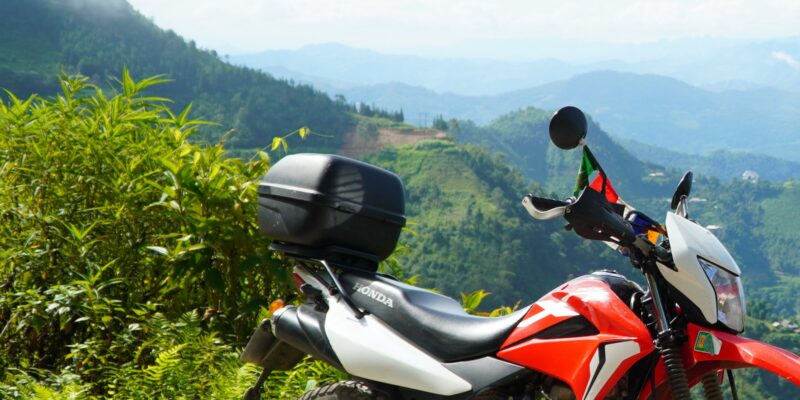 A red trail motorbike parked on a gravel shoulder above terraced fields, a top box on the rack, with green ridges and low cloud behind