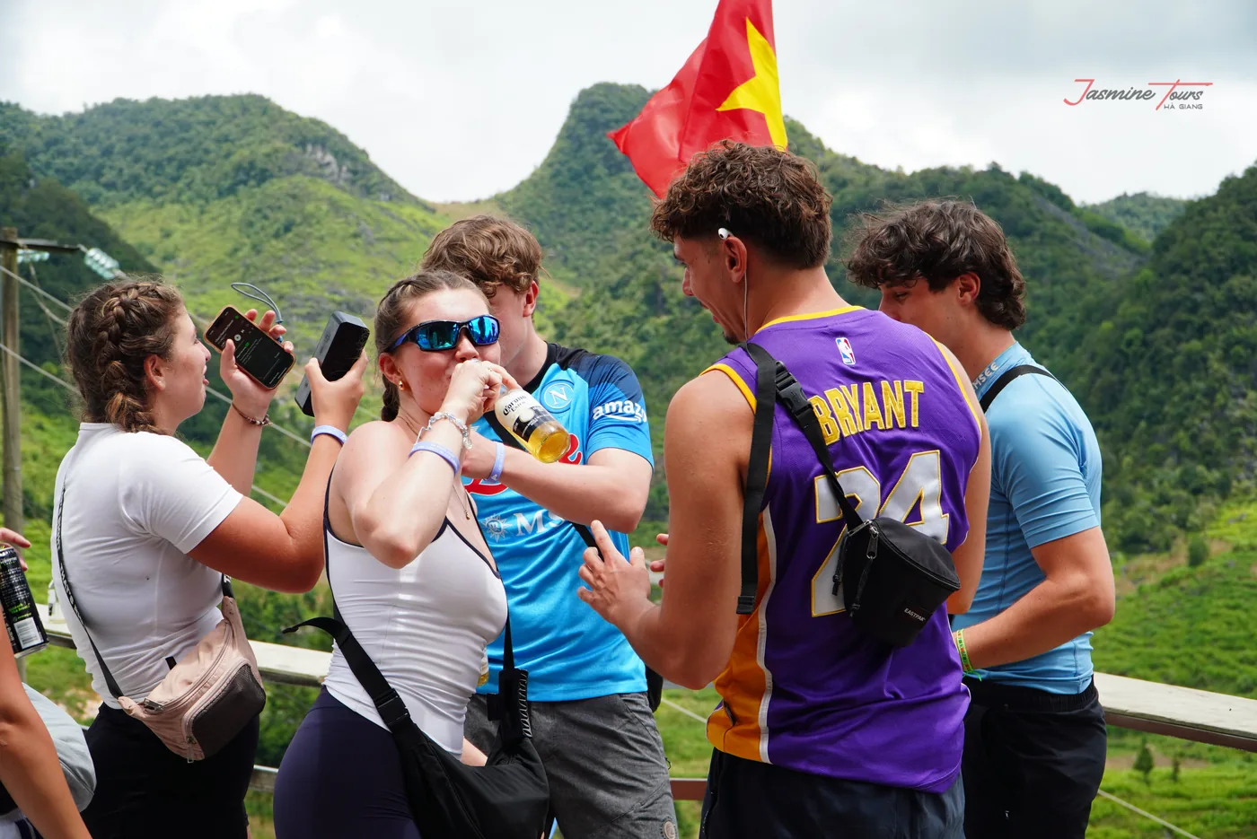 Three travellers at a roadside viewpoint, one photographing the others against a valley of green karst ridges, a red flag on a pole beside them and a road visible on the far slope