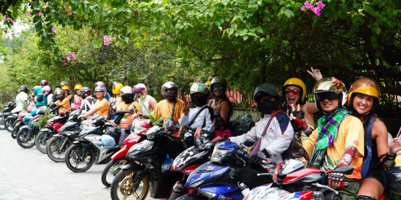A long line of small motorbikes parked nose-in along a roadside, more than a dozen of them, riders in helmets and orange jerseys sitting on or standing beside them, green hillside behind and a strip of cloud-covered ridge above