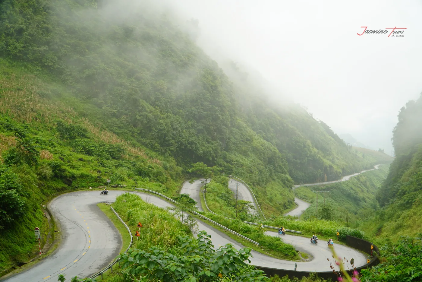 A road folding back on itself through four visible hairpins on a green mountainside, the upper half of the valley lost in low white cloud, and two motorbikes small in the frame near the lower bends