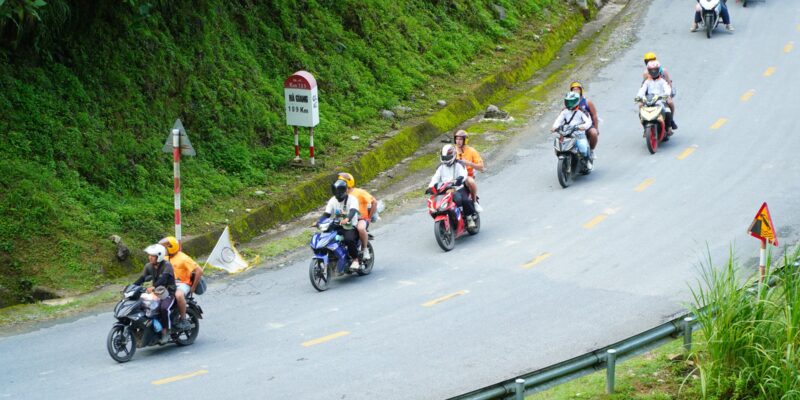 A loose column of six small motorbikes coming down a two-lane mountain road, two riders in orange jerseys carrying pillion passengers at the front of the group, a red-and-white kilometre marker at the roadside reading QL.4C Km 109 and HA GIANG 109 Km, a steel guardrail along the outer edge and steep green vegetation on the uphill side