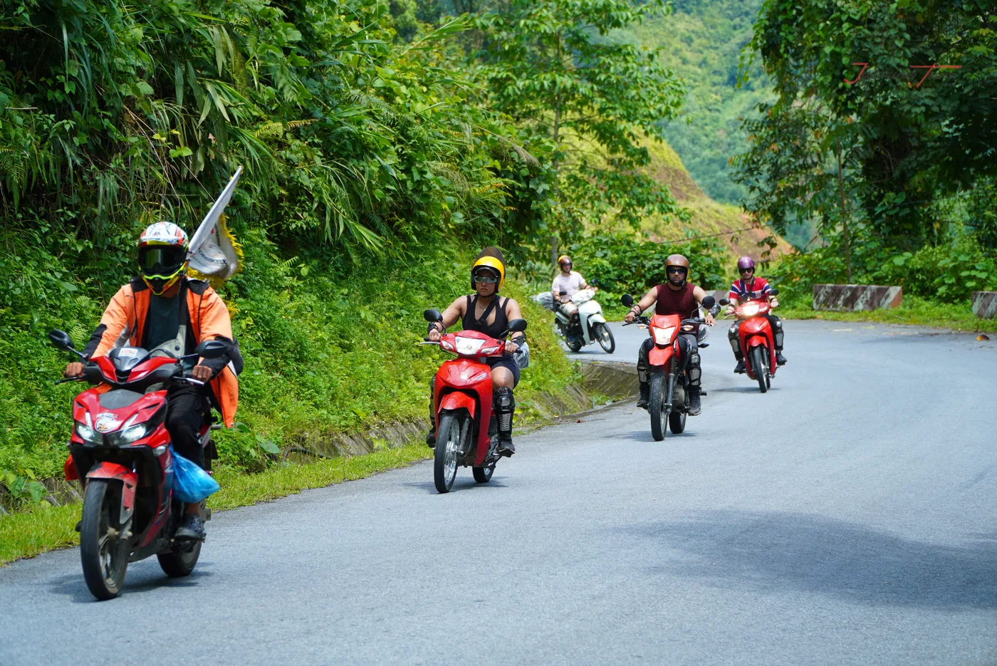 A guide in an orange jersey riding at the front of a loose column on a two-lane mountain road, a small white flag mounted on the back of his bike, four more riders strung out behind him, thick green vegetation on the uphill side and a concrete drainage channel along the verge