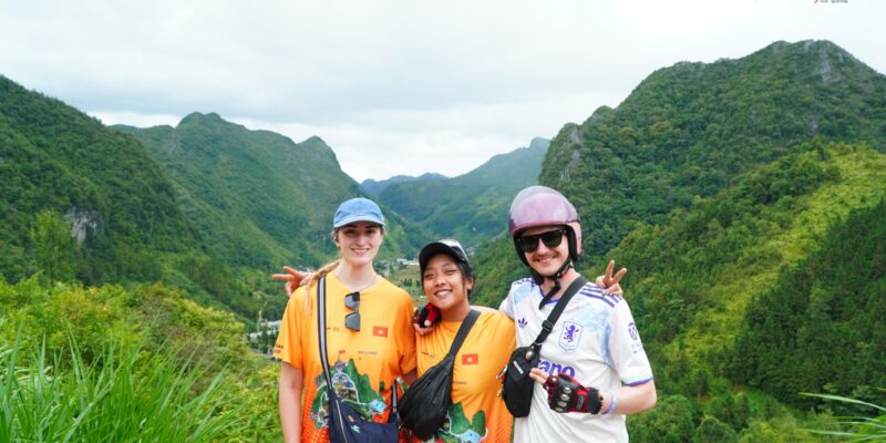 Three travellers in helmets standing on a grassy roadside bank with green karst ridges layered behind them
