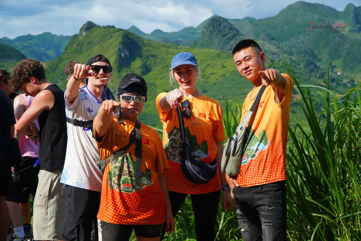 A group of travellers standing together at a roadside viewpoint with arms round each other, steep grey rock rising behind them and green vegetation along the verge