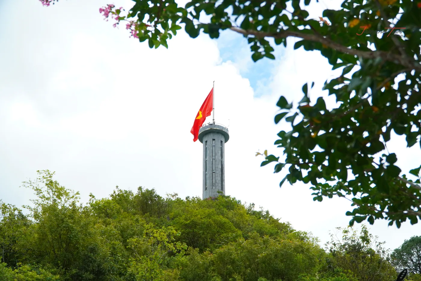 A stone flag tower on a wooded hilltop seen from below through the branches of a flowering tree, with a railed viewing gallery near the top and a large red flag with a yellow star flying from the mast above it against a pale overcast sky