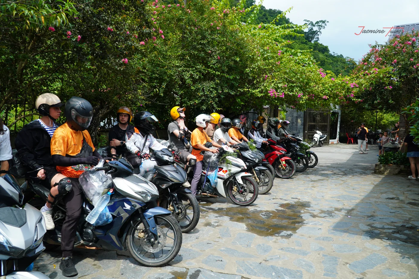A line of small motorbikes parked nose-in along a wet flagstone path under flowering shrubs, a dozen riders sitting on or standing beside them, every one of them in a helmet, with knee pads and rain covers over the luggage
